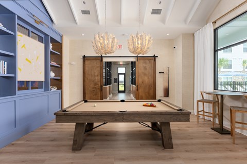 A pool table is in the middle of a room with a blue wall and a chandelier hanging from the ceiling. at Evolve Holly Ridge Apartments in Holly Ridge, NC