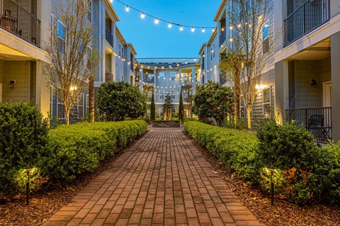 A long brick pathway leads through a well-manicured garden and between apartment buildings.
