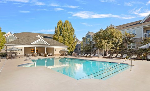 A large swimming pool in front of a house with a patio and lounge chairs.