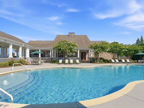 A large swimming pool in front of a house with a patio and lounge chairs.