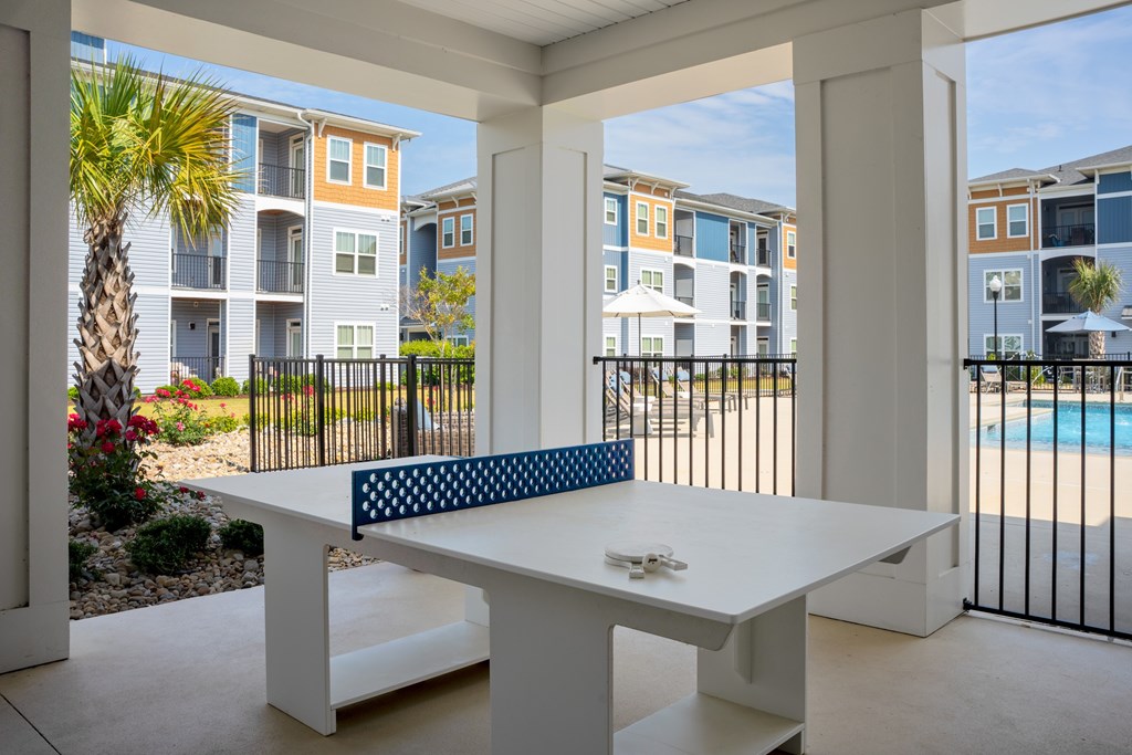 A white table with a blue cushion is in the foreground of a patio with a pool and apartment buildings in the background.