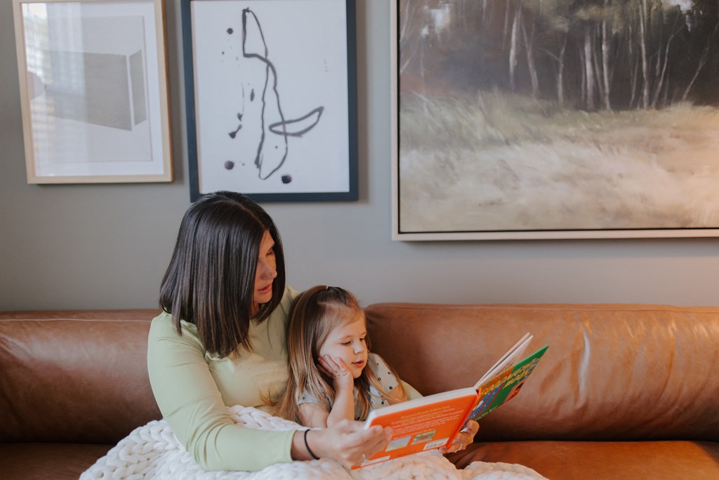 A woman and a child are sitting on a brown couch reading a book.