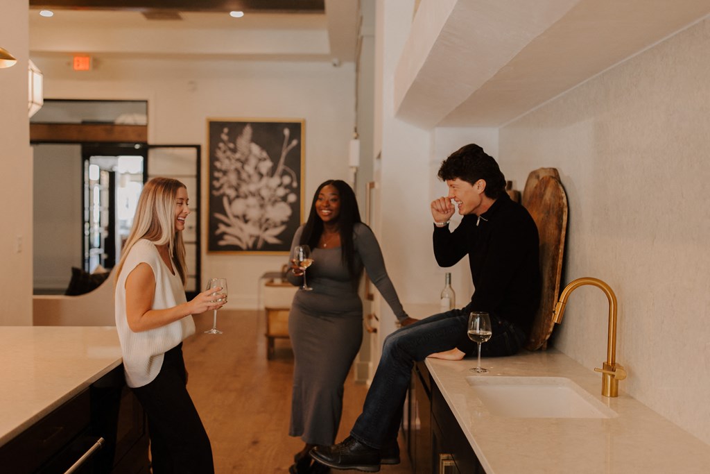 Three people are standing in a kitchen, two of them are holding wine glasses.
