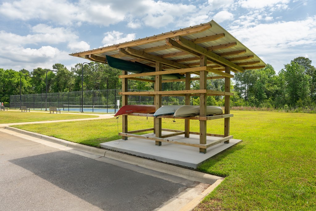 A wooden pavilion with a red roof is situated in a grassy area next to a paved road.