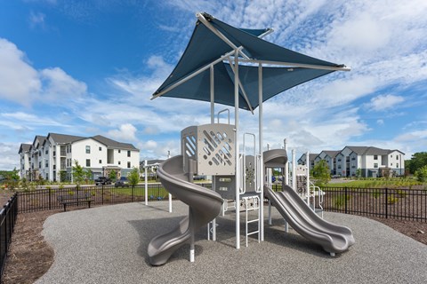 A playground with a slide and a climbing structure under a blue umbrella at Evolve Holly Ridge Apartments in Holly Ridge, NC.