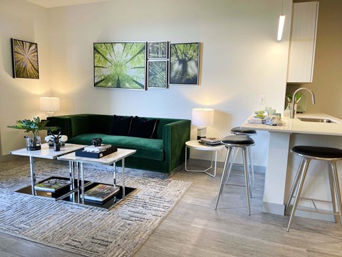 A living room with a green couch, a white coffee table, and a kitchen area with a sink and bar stools.