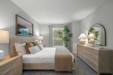 A well-lit bedroom with a large bed, a nightstand with a lamp, and a mirror on the wall.