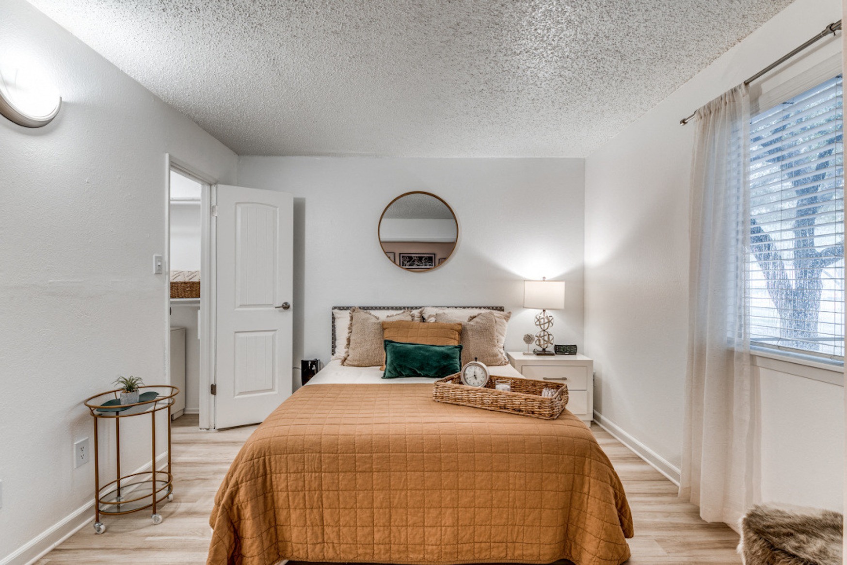 Renovated bedroom in Fusion Fort Worth, previously Desert Sands apartments. Large bed with camel duvet and green velvet pillow next to midcentury modern bar cart with succulent.