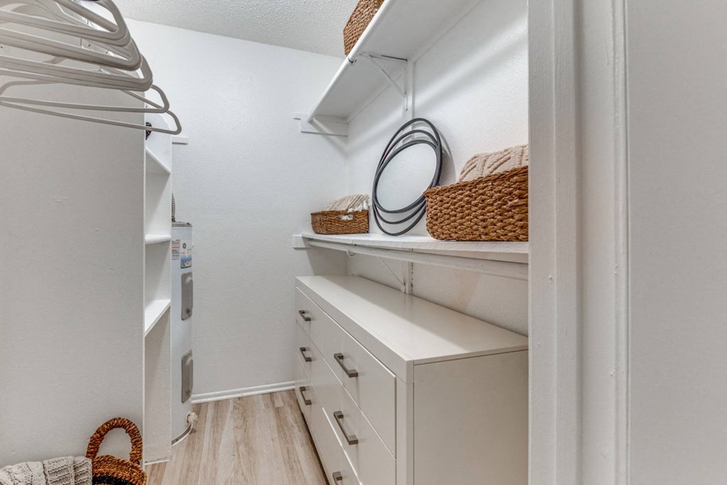 Fusion Fort Worth apartment walk in closet with wicker baskets on built in shelving, hangers on curtain rods, and white midcentury modern dresser.