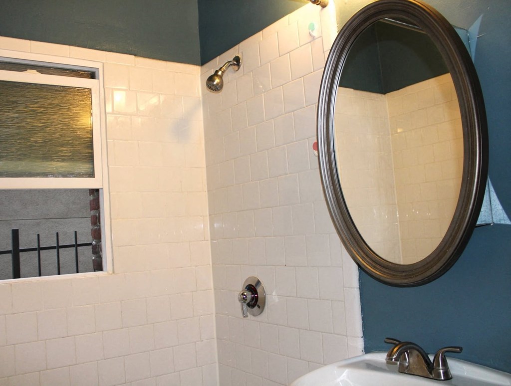 Bathroom in one bedroom brookmore apartment in Pasadena. Shower with white tile and silver fixtures and open window, and oval mirror above pedestal sink