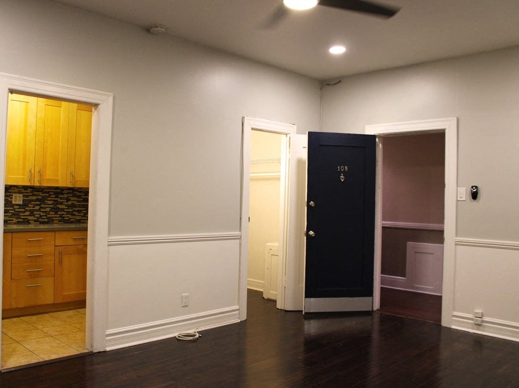 Living room in brookmore apartments in Pasadena featuring dark wood flooring and white wainscoting. Open doorways lead to the kitchen and front closet.