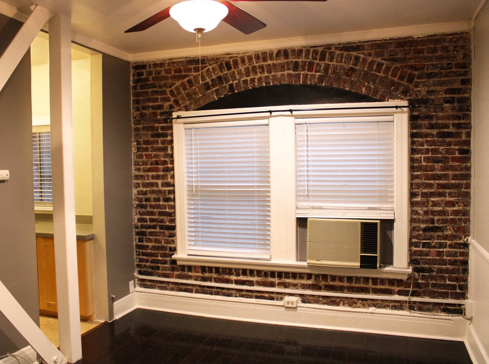 Living room in brookmore apartments in Pasadena featuring an exposed brick wall, large windows with shades, and a window air conditioning unit.