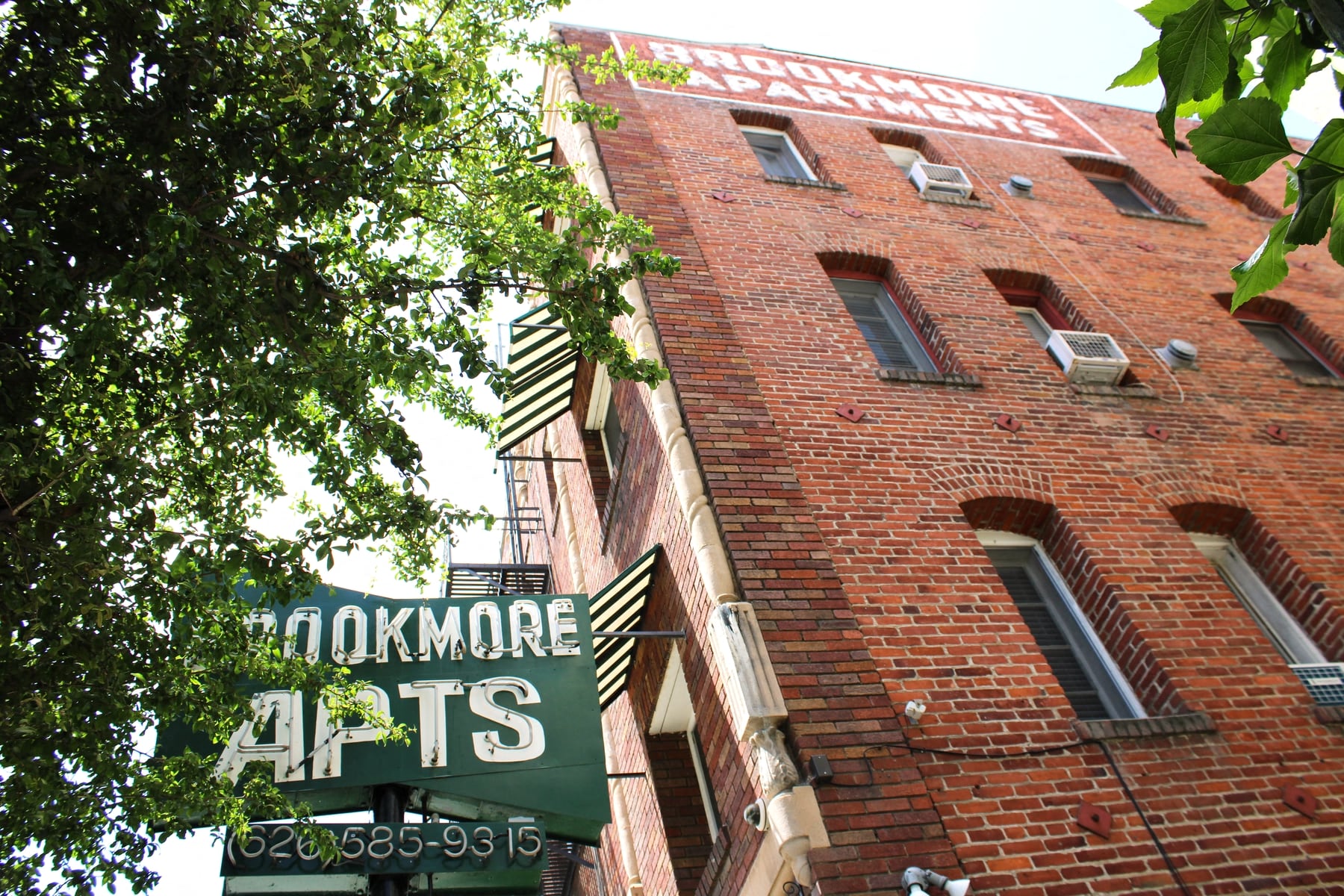 Upshot of large brick building in Pasadena CA. Two large signs saying "Brookmore apts" and "Brookmore Apartments" on building with green trees in front.