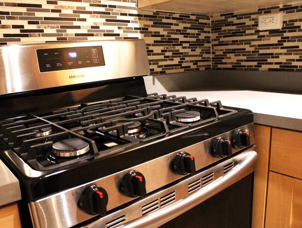 A six burner gas stove in a brookmore apartments kitchen with honey colored wood cabinets and multicolored backsplash.