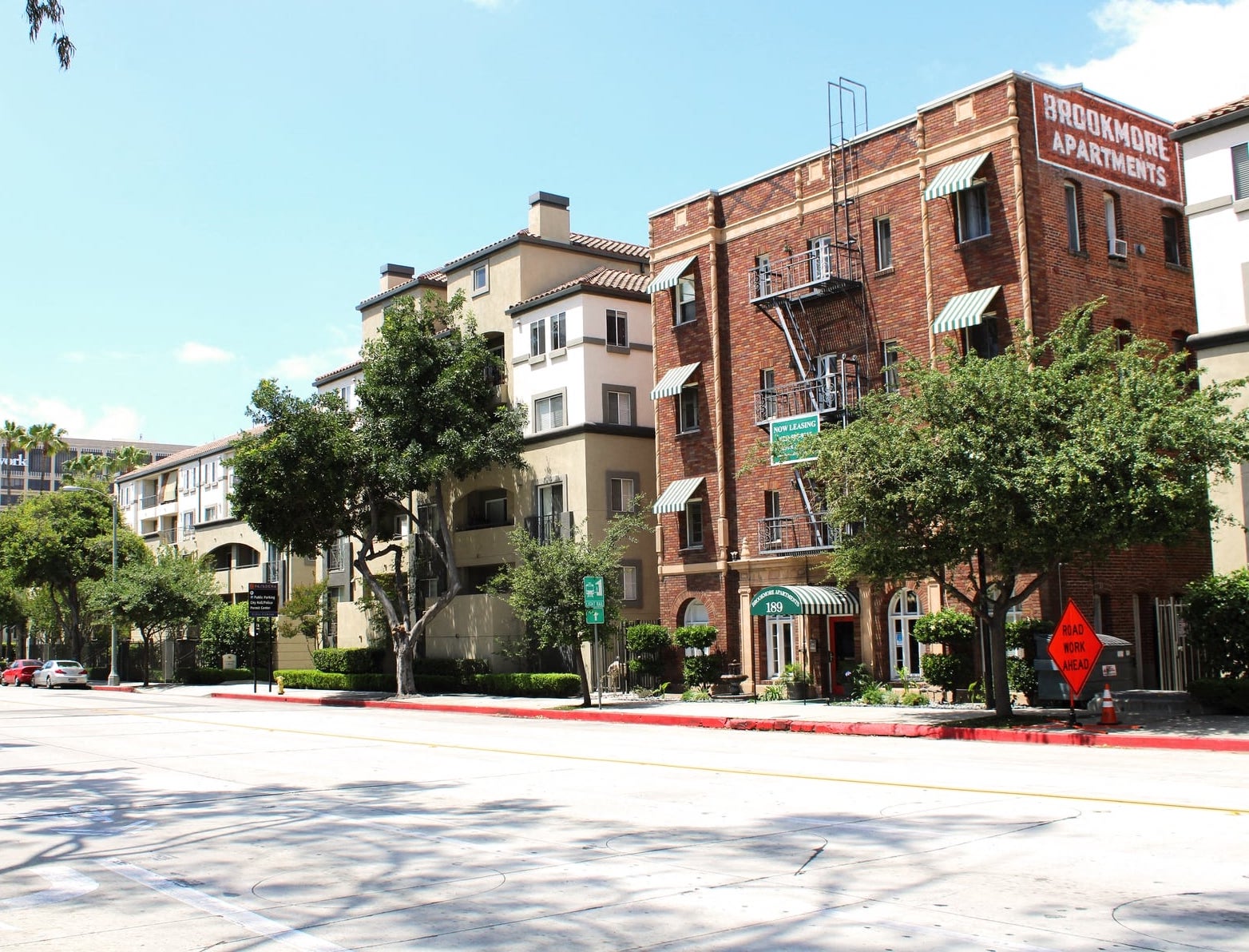 Street view of Brookmore apts in Pasadena CA. Four story brick building with "now leasing" sign and large green trees and bushes in front