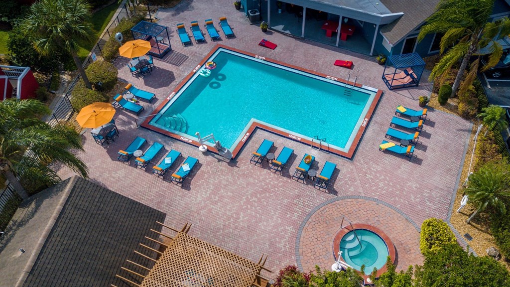 Community pool at Fusion Orlando Apartments aerial shot. Pool is surrounded by sixteen blue lounge chairs and patio tables with orange umbrellas.