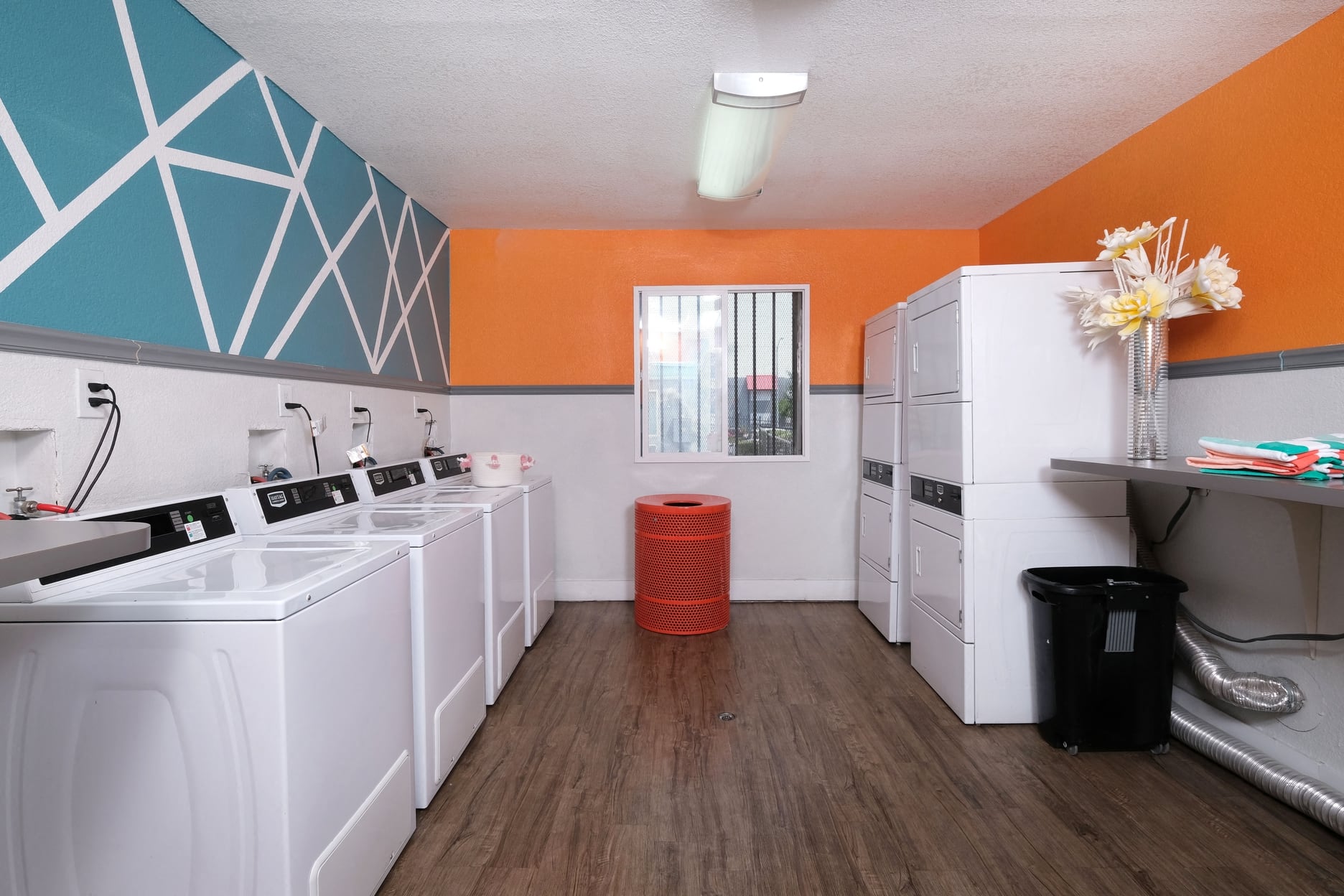 On site laundry at Fifteen 50 apartments in Las Vegas with four washers and dryers, red trash can, and modern orange, blue, and white walls.