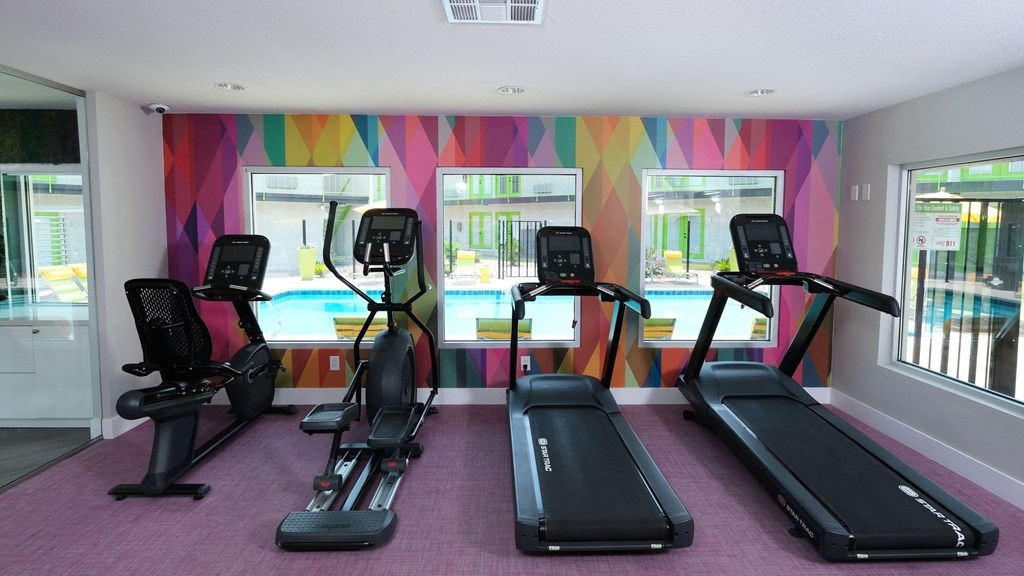 Cardio machines looking out three large windows in a colorful wall in the fitness center at Fusion Las Vegas Apartments.