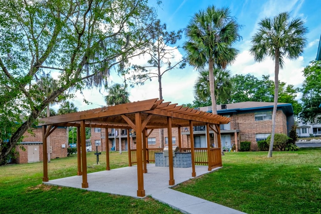 BBQ area at Watermarc Apartments in Lakeland, FL with stained wood trellis on concrete slab surrounded by green grass and palm trees.