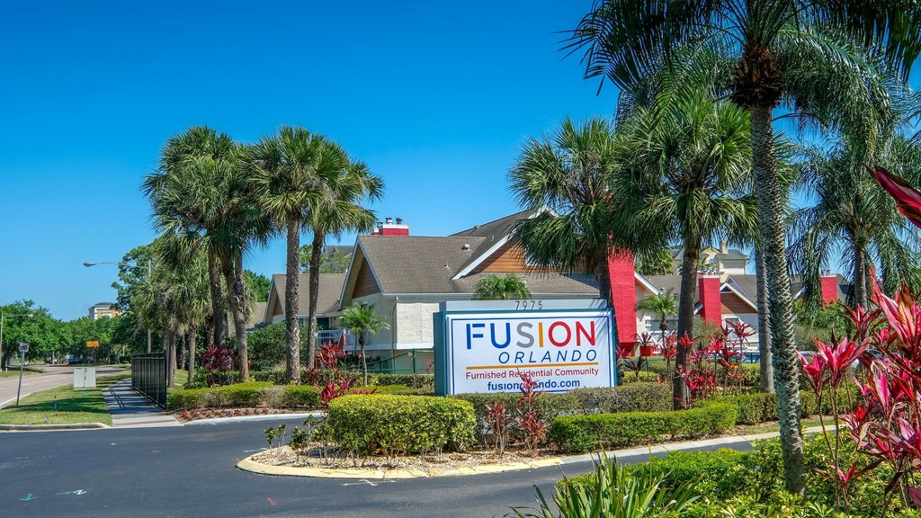 Main entrance to Fusion furnished apartments in Orlando, FL. Palm trees, bushes, and tropical flora line the main road and median with Fusion Orlando sign.