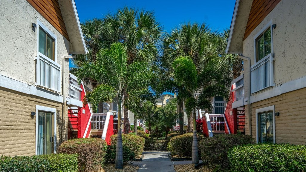 Fusion Orlando apartments pathway between buildings lined with lush bushes and palm trees. Red staircases lead up to second story.