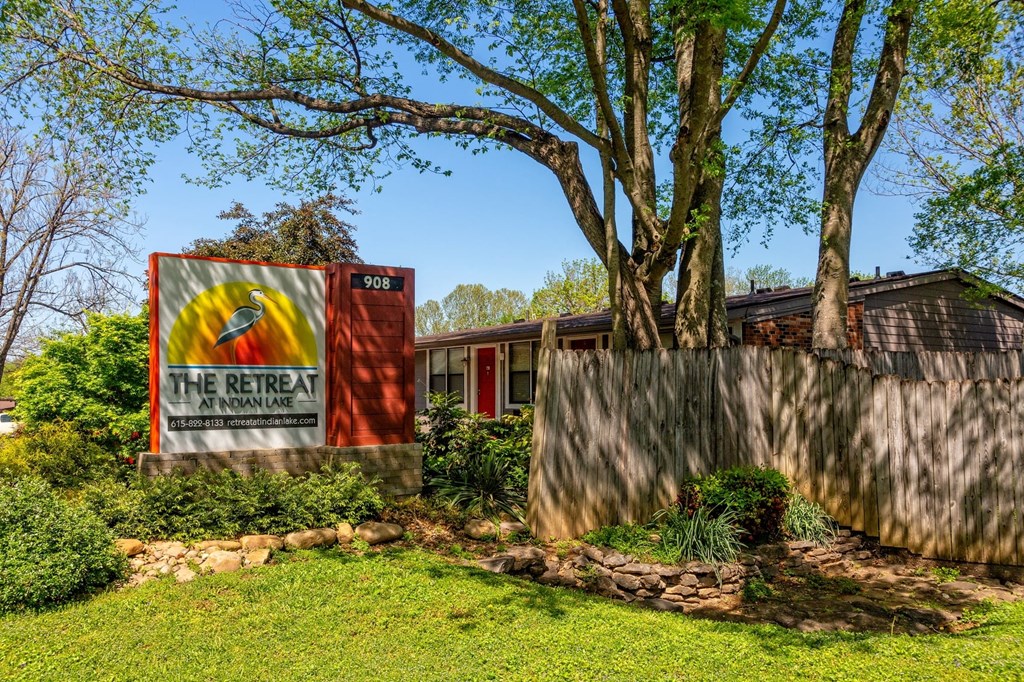 The Retreat at Indian Lake apartments monument sign in river rock planter with green landscaped bushes and shaded by tall trees.