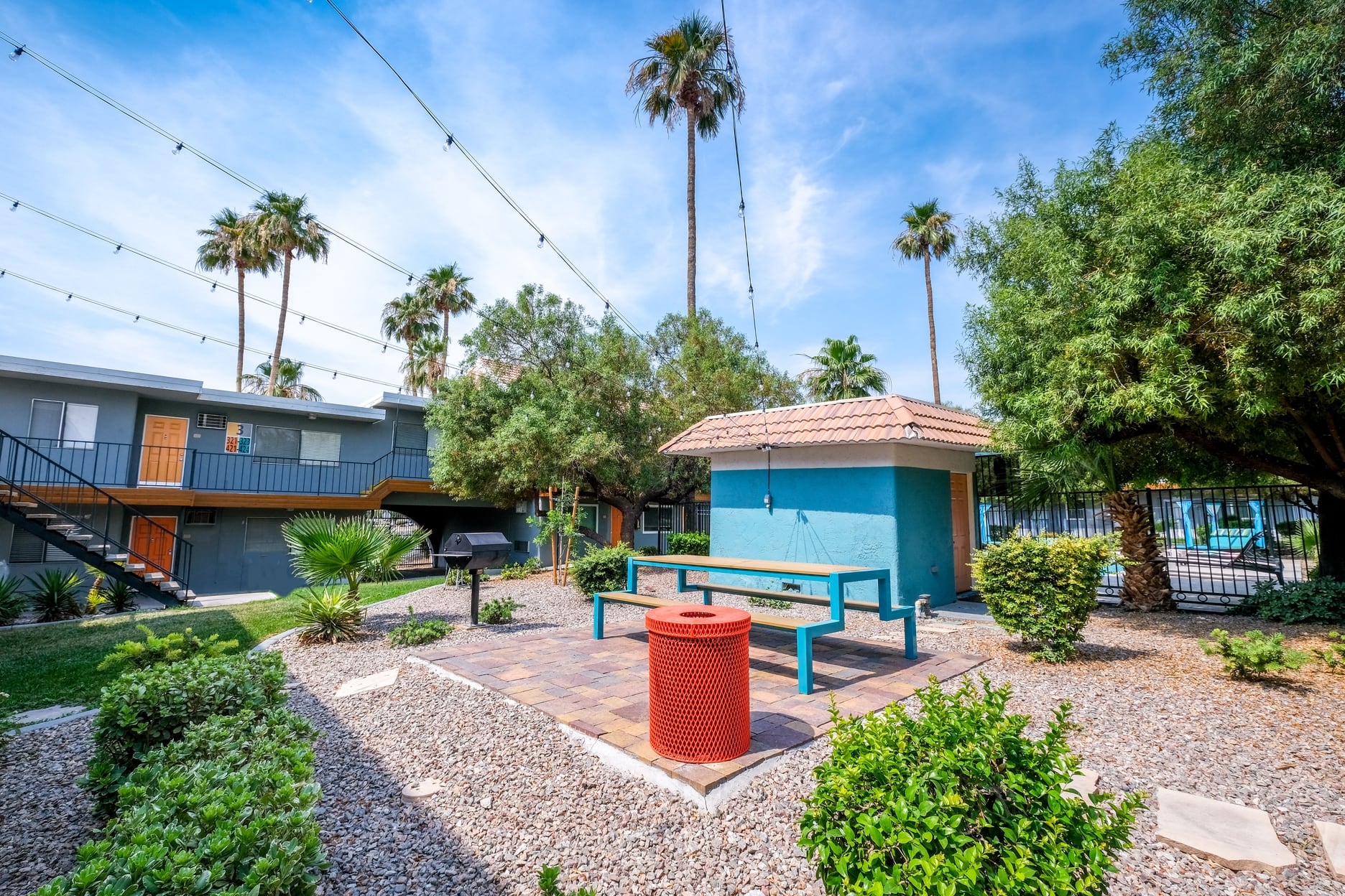 Fifteen 50 apartments Las Vegas courtyard with modern blue picnic table, market lights, red trash can, and barbecue next to small blue structure.