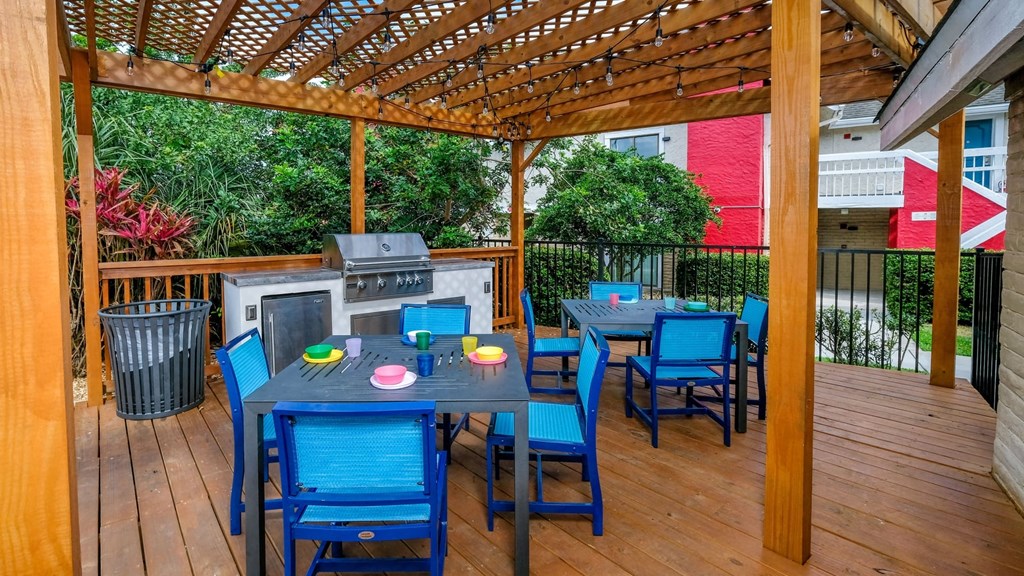 Fusion Orlando apartments outdoor grilling area with tables and chairs, grill and prep counters, and trash can under stained wood trellis.