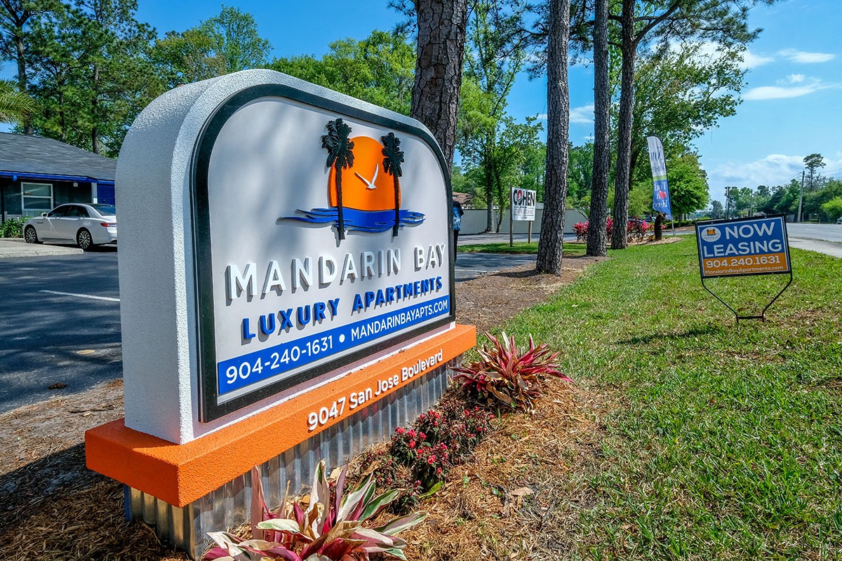 Street sign in Jacksonville, Florida that reads "Mandarin Bay Luxury Apartments" next to row of tall trees and "now leasing" sign.