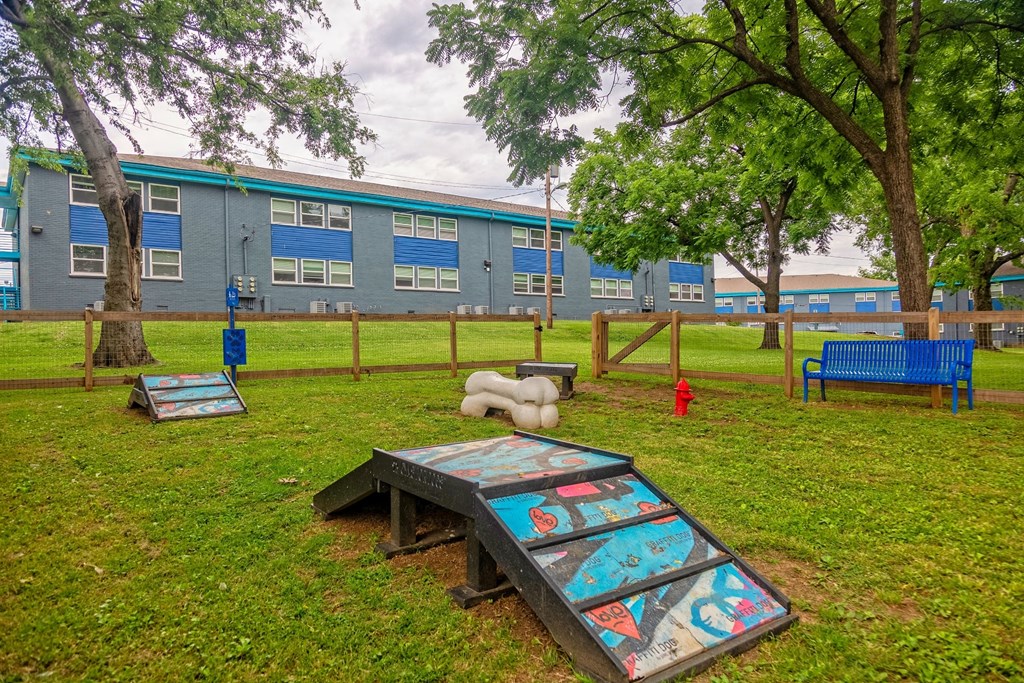 The Canvas apartments dog park with agility course, pet waste station, large green trees, and apartment buildings in the background.