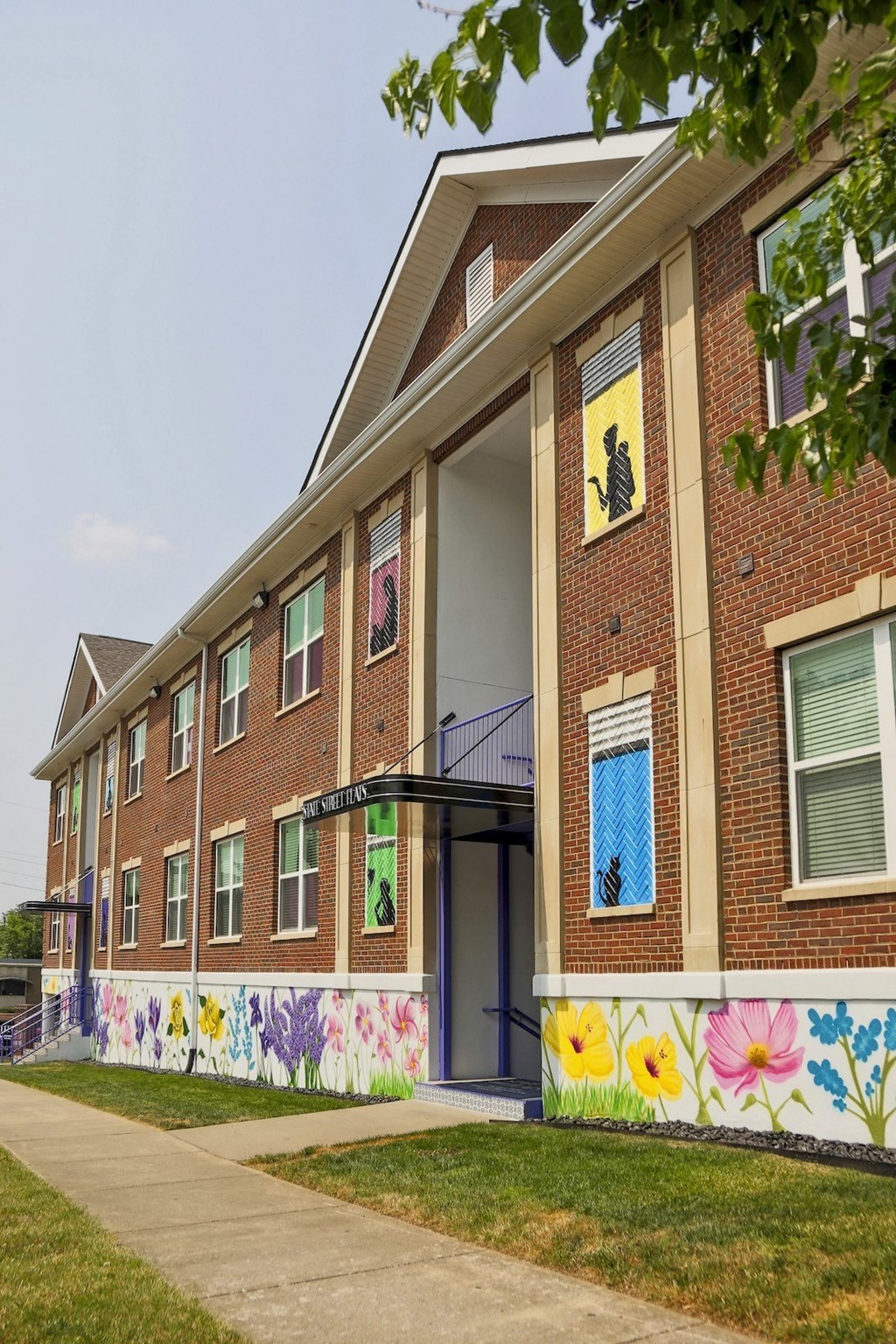State Street Flats Apartment building exterior with two entrances to brick building with floral mural and green grass.