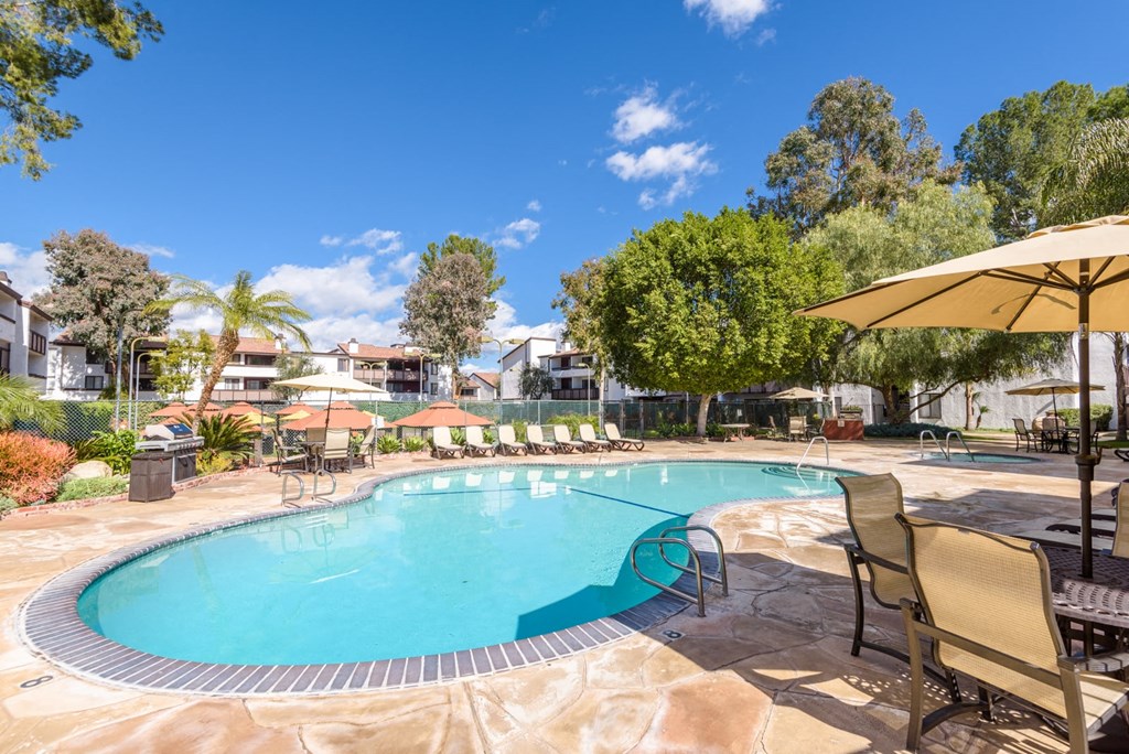 Fusion Warner Center apartments in Woodland Hills pool with tan lounge chairs and patio tables with tan umbrellas on natural stone tile ground.