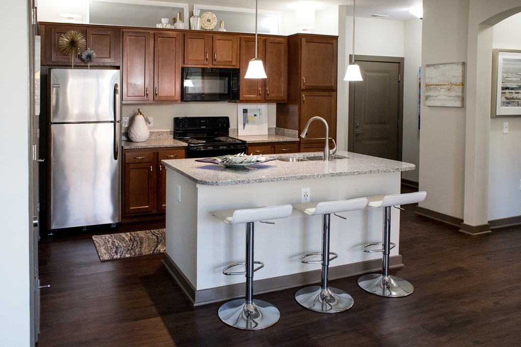 One BR Apartments in Miamisburg, OH - Austin Park - Kitchen with Dark Brown Cabinets, Granite-Style Countertops, Island with Barstools, and Stainless Steel Appliances.
