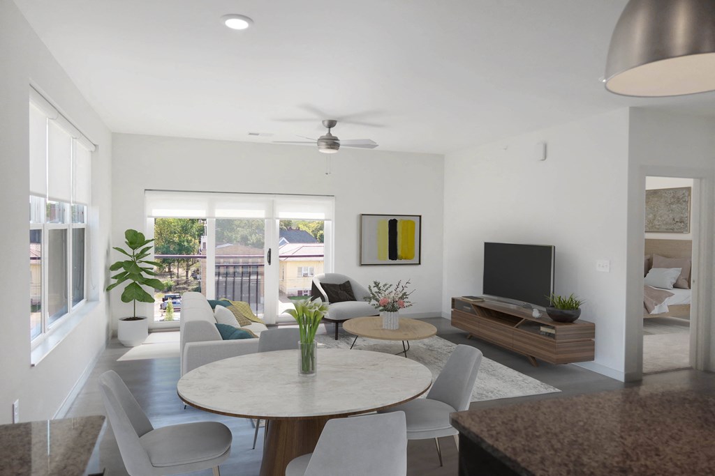 A dining room table surrounded by four chairs, next to a furnished living room with a ceiling fan and a sliding glass door.