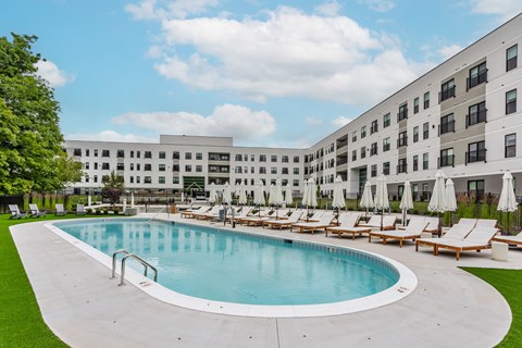 A large swimming pool surrounded by sun loungers and umbrellas in front of a modern building.