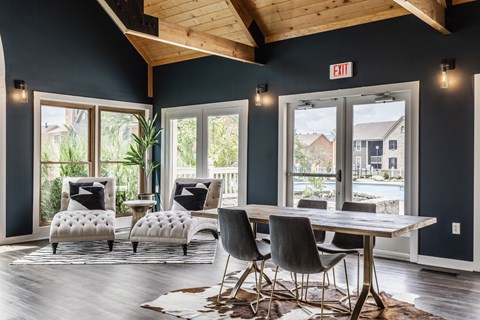 A community clubhouse featuring two cozy comfy chairs, a table surrounded by chairs, hardwood-style flooring, and large windows.