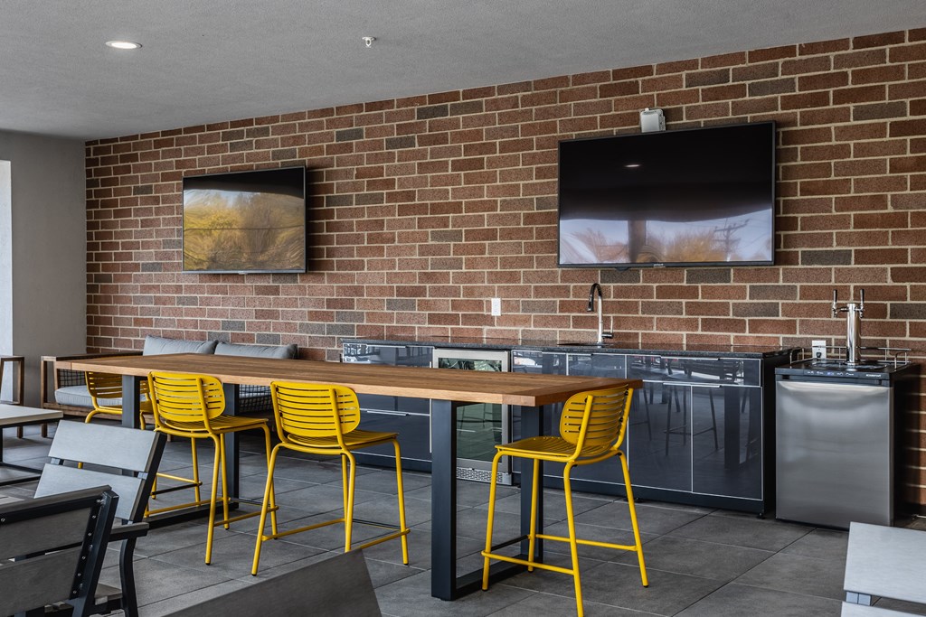 A sky lounge with a large table surrounded by four yellow chairs, next to two TV's on a brick wall.
