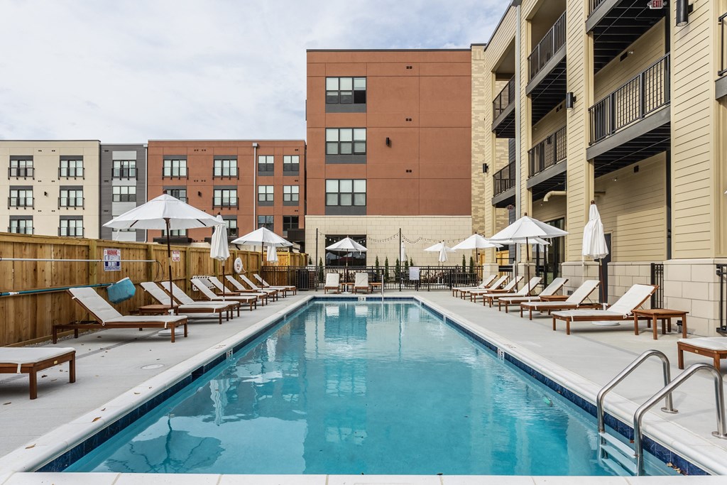 A swimming pool surrounded by a fence, white chairs and white umbrellas, next to apartment buildings.