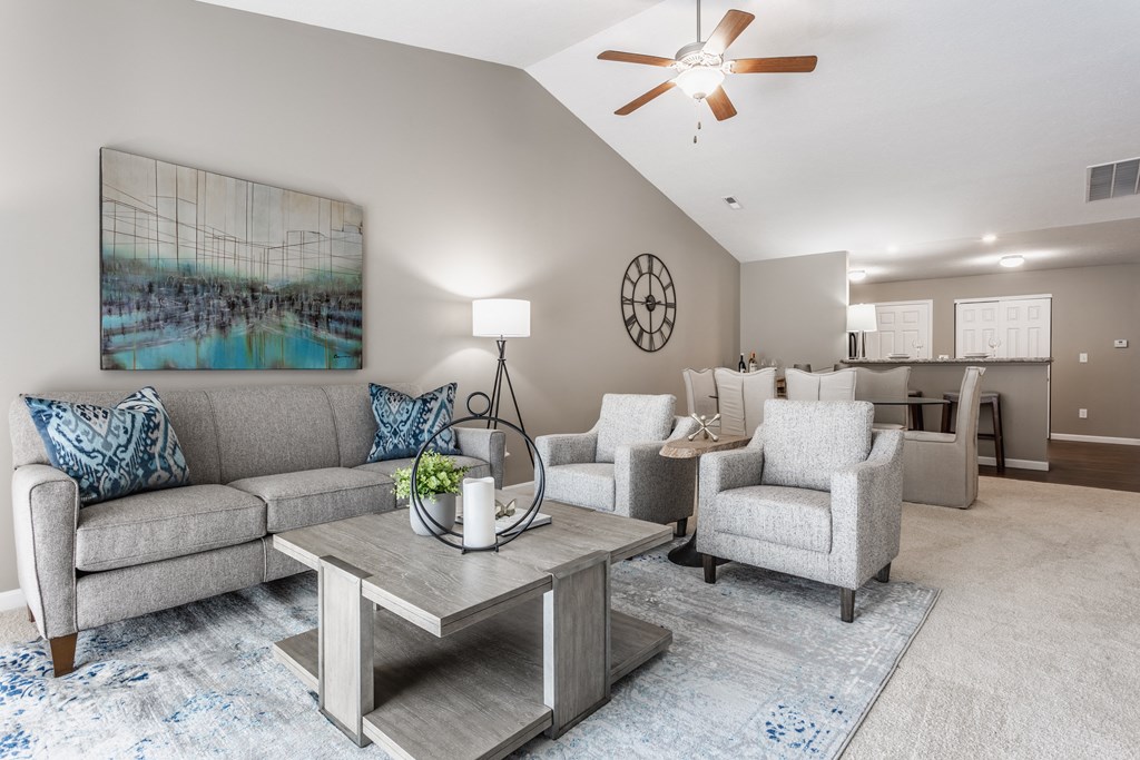 Wheaton Village living room with a grey couch and two grey chairs, a center table, and carpeted floors, next to a dining room table.