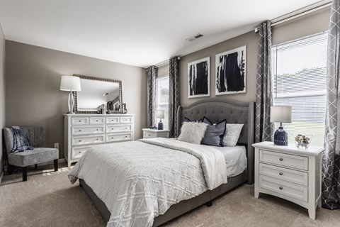 A furnished bedroom with carpeted flooring, stylish decor, and two windows for natural light.