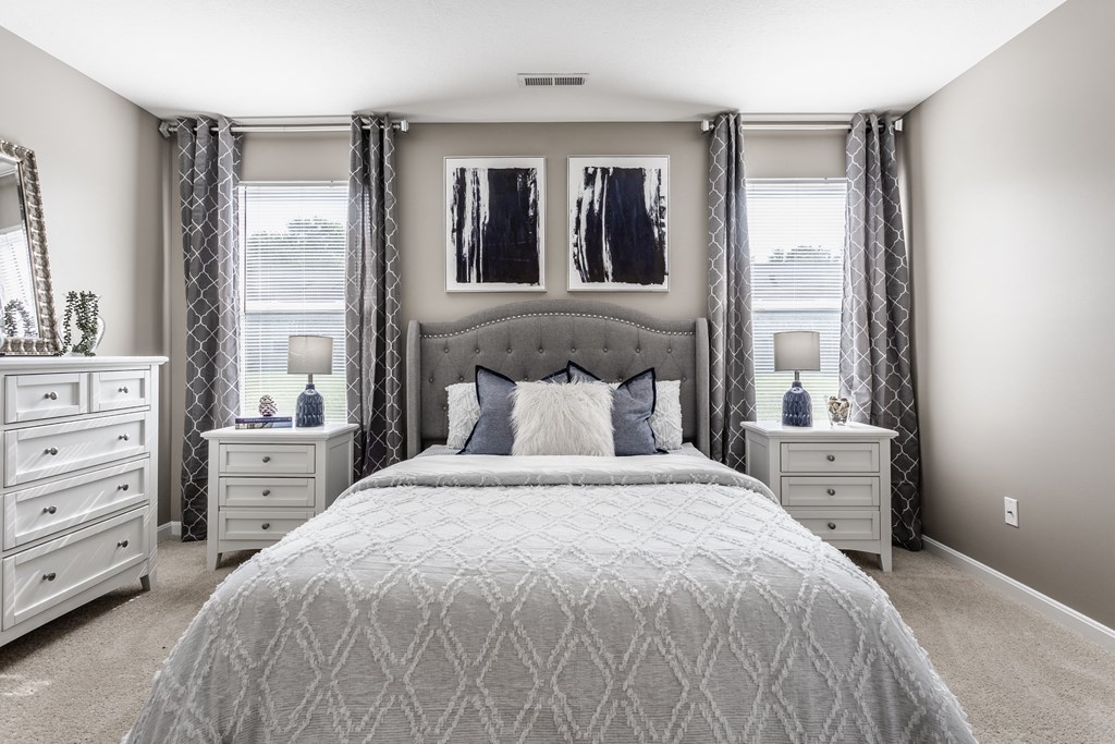 A bedroom with a bed, two bedside tables each with a lamp, a dresser, and two windows for natural light.