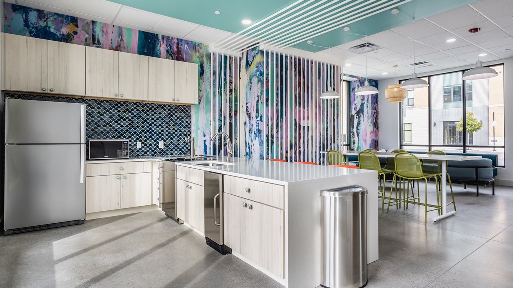 A lounge with a kitchen with stainless steel appliances, a light wood cabinets, next to a table and chairs.