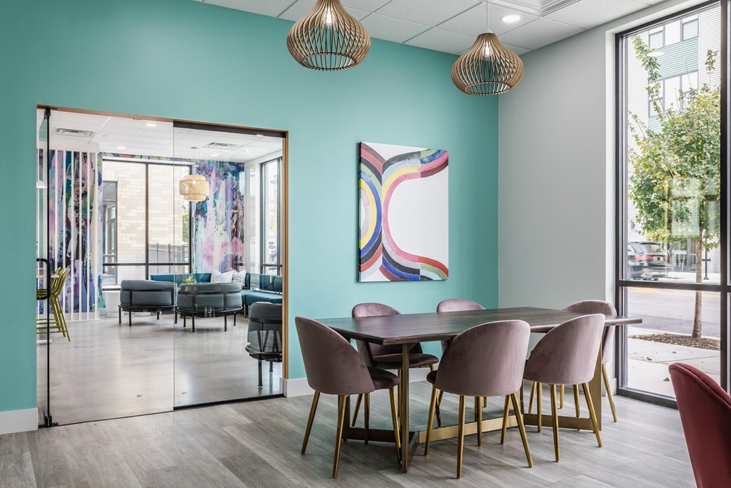 A co-working lounge at Madamore with a blue wall, a table surrounded by six chairs, and a large window, on hardwood-style floors.