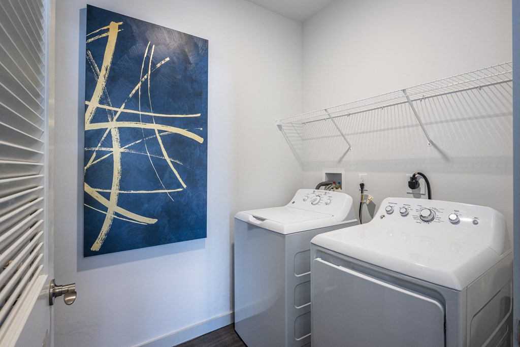 A closet with a shelf, a white washer and dryer, and a painting on the wall.