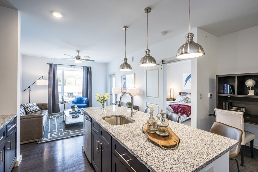 Kitchen featuring an island with a sink and granite countertops, next to a living room with a large window.