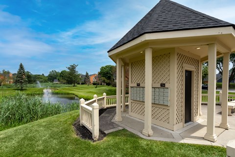 A gazebo with a black roof is situated in a grassy area with a pond and trees in the background.