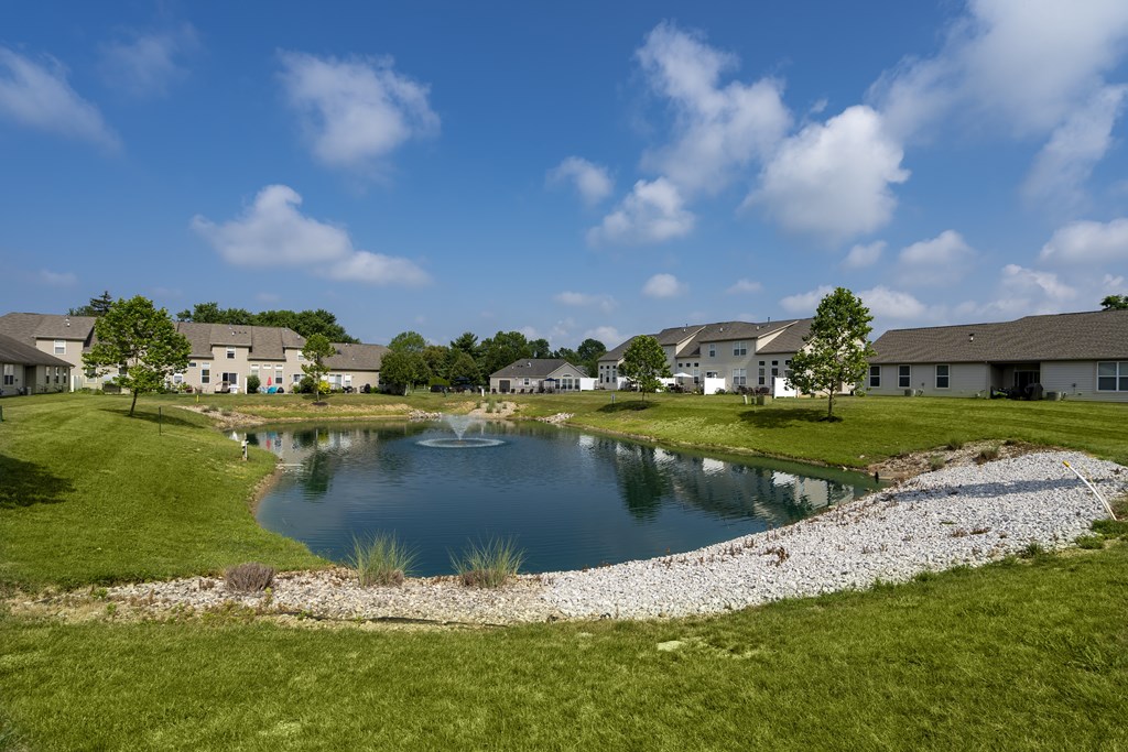 A pond in the middle of a grassy area with houses in the background.