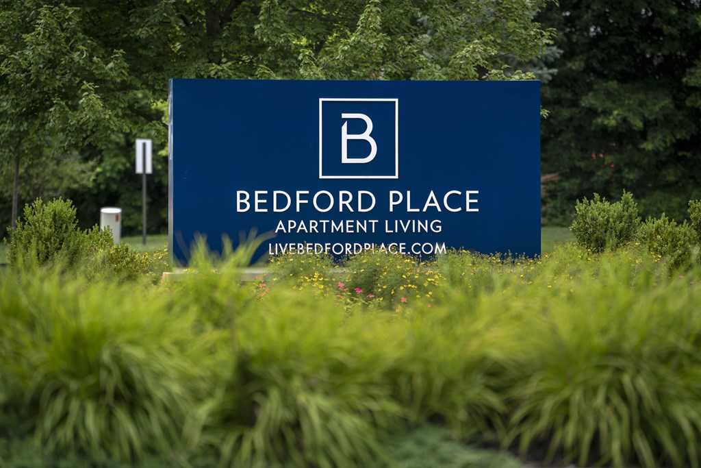 A blue sign for Bedford Place Apartment Living in front of some greenery.