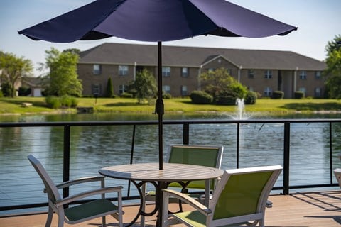 A table with a purple umbrella is set up on a deck overlooking a pond.