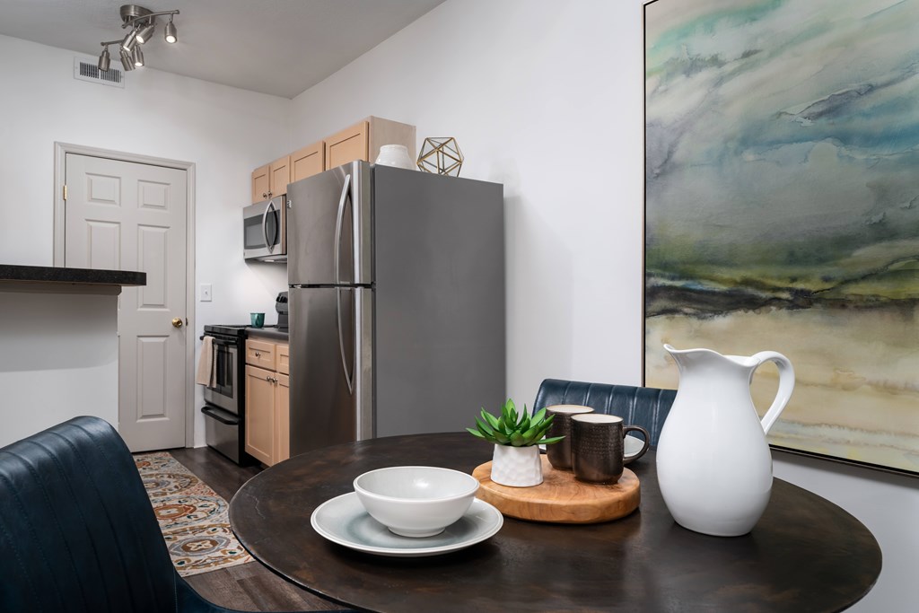 A modern kitchen with a refrigerator, a dining table with a bowl, a plant, and a jug.